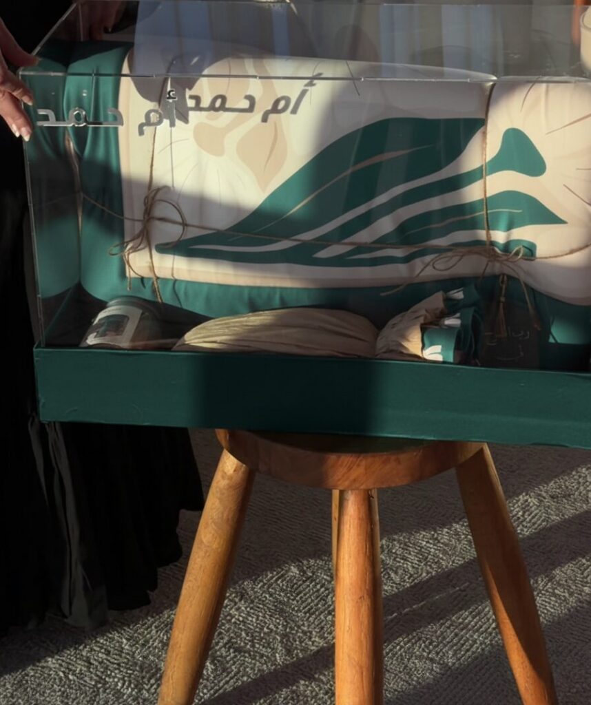 نقصة المحبة الخضراء معروضة في صندوق أكريليك على طاولة خشبية مع سجادة الصلاة الخضراء وبطاقة اسم