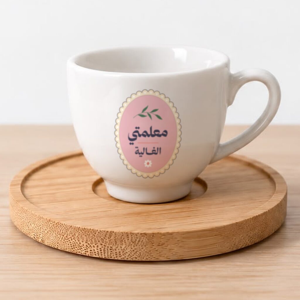 فنجان قهوة سيراميك أبيض عليه رسمة غصن أخضر وزهرة صغيرة على خلفية وردية وعبارة «معلمتي الغالية»، على قاعدة خشبية