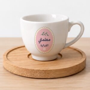 فنجان قهوة سيراميك أبيض عليه رسمة غصن أخضر وزهرة صغيرة على خلفية وردية وعبارة «معلمتي الغالية»، على قاعدة خشبية