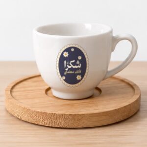 فنجان قهوة سيراميك أبيض عليه رسمة أزهار أقحوان صغيرة على خلفية كحلية وعبارة «شكرًا لأنكِ معلمتي»، على قاعدة خشبية