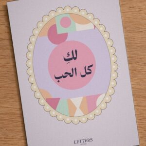 بطاقة إهداء بمقاس A5 عليها رسمة أشكال هندسية بالوردي والبنفسجي وعبارة «لكِ كل الحب»، على طاولة خشبية