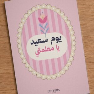 بطاقة إهداء بمقاس A5 عليها رسمة زهرة توليب على خطوط وردية وعبارة «يوم سعيد يا معلمتي»، على طاولة خشبية