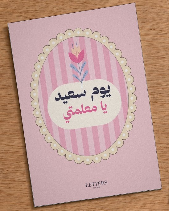 بطاقة إهداء بمقاس A5 عليها رسمة زهرة توليب على خطوط وردية وعبارة «يوم سعيد يا معلمتي»، على طاولة خشبية