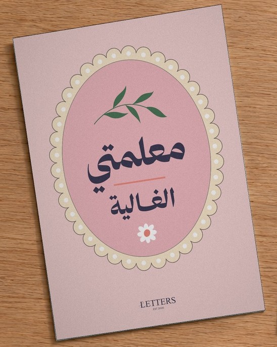 بطاقة إهداء بمقاس A5 عليها رسمة غصن أخضر وزهرة صغيرة على خلفية وردية وعبارة «معلمتي الغالية»، على طاولة خشبية