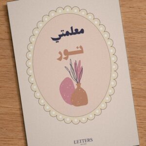 بطاقة إهداء بمقاس A5 عليها رسمة مزهرية بأغصان مجففة على خلفية وردية فاتحة وعبارة «معلمتي نور»، على طاولة خشبية