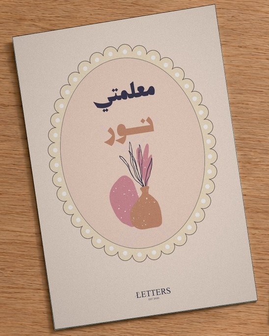 بطاقة إهداء بمقاس A5 عليها رسمة مزهرية بأغصان مجففة على خلفية وردية فاتحة وعبارة «معلمتي نور»، على طاولة خشبية