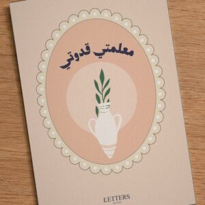بطاقة إهداء بمقاس A5 عليها رسمة جرّة فخارية بغصن أخضر على خلفية خوخية وعبارة «معلمتي قدوتي»، على طاولة خشبية