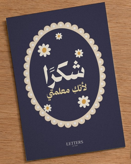 بطاقة إهداء بمقاس A5 عليها رسمة أزهار أقحوان صغيرة على خلفية كحلية وعبارة «شكرًا لأنكِ معلمتي»، على طاولة خشبية