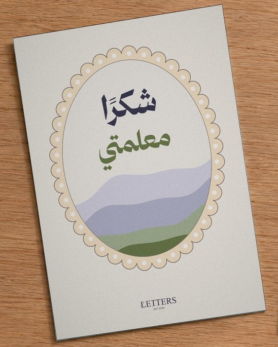 بطاقة إهداء بمقاس A5 عليها رسمة تلال خضراء وبنفسجية وعبارة «شكرًا معلمتي»، على طاولة خشبية