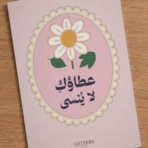 بطاقة إهداء بمقاس A5 عليها رسمة زهرة أقحوان على خلفية وردية وعبارة «عطاؤكِ لا يُنسى»، على طاولة خشبية