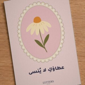 بطاقة إهداء بمقاس A5 عليها رسمة زهرة كبيرة كريمية بقلب برتقالي على خلفية وردية وعبارة «عطاؤكِ لا يُنسى»، على طاولة خشبية