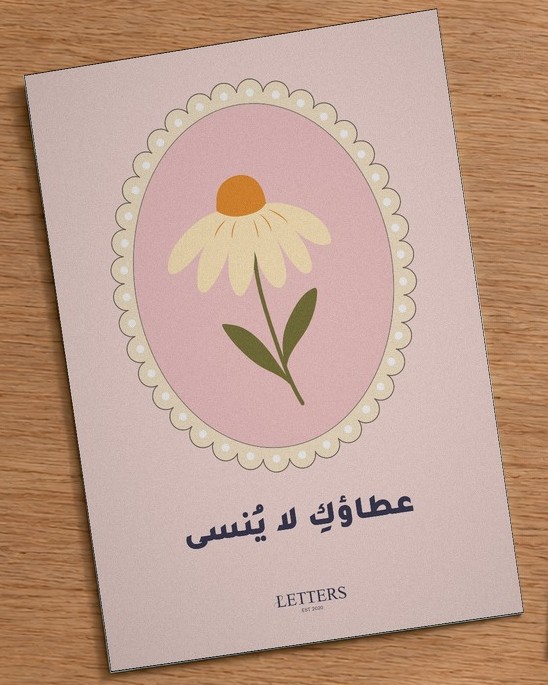 بطاقة إهداء بمقاس A5 عليها رسمة زهرة كبيرة كريمية بقلب برتقالي على خلفية وردية وعبارة «عطاؤكِ لا يُنسى»، على طاولة خشبية