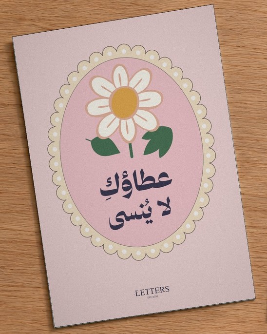 بطاقة إهداء بمقاس A5 عليها رسمة زهرة أقحوان على خلفية وردية وعبارة «عطاؤكِ لا يُنسى»، على طاولة خشبية