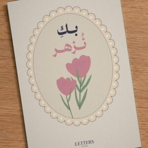 بطاقة إهداء بمقاس A5 عليها رسمة أزهار وردية على خلفية فاتحة وعبارة «بكِ نُزهر»، على طاولة خشبية