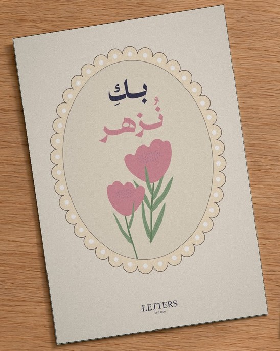 بطاقة إهداء بمقاس A5 عليها رسمة أزهار وردية على خلفية فاتحة وعبارة «بكِ نُزهر»، على طاولة خشبية