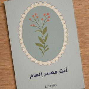 بطاقة إهداء بمقاس A5 عليها رسمة غصن بأزهار حمراء صغيرة على خلفية خضراء رمادية وعبارة «أنتِ مصدر إلهام»، على طاولة خشبية