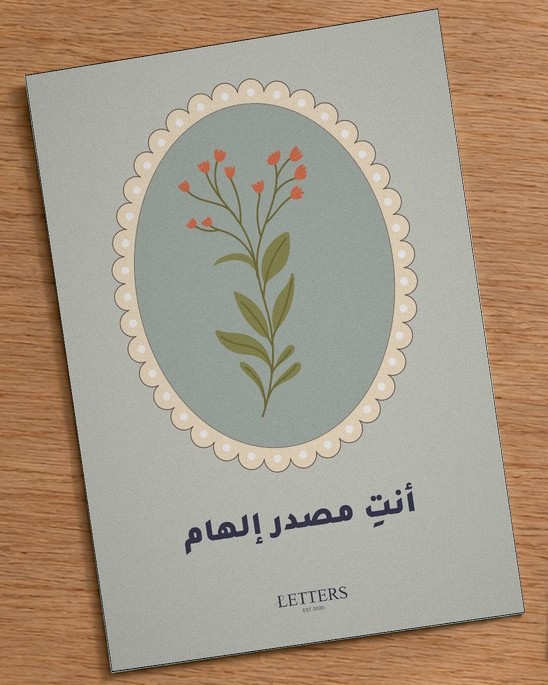 بطاقة إهداء بمقاس A5 عليها رسمة غصن بأزهار حمراء صغيرة على خلفية خضراء رمادية وعبارة «أنتِ مصدر إلهام»، على طاولة خشبية