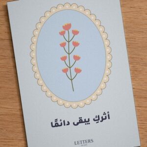 بطاقة إهداء بمقاس A5 عليها رسمة أزهار وردية صغيرة على غصن فوق خلفية زرقاء فاتحة وعبارة «أثركِ يبقى دائمًا»، على طاولة خشبية