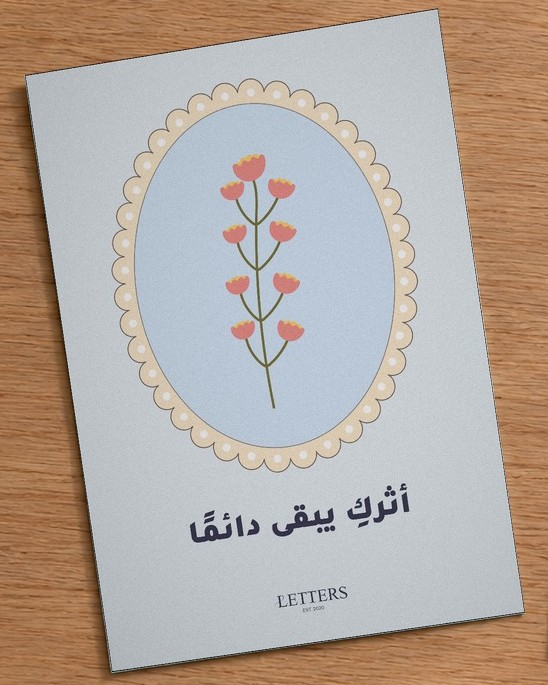 بطاقة إهداء بمقاس A5 عليها رسمة أزهار وردية صغيرة على غصن فوق خلفية زرقاء فاتحة وعبارة «أثركِ يبقى دائمًا»، على طاولة خشبية